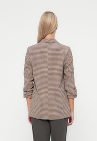Blazer à carreaux marron avec un col montant et des manches trois-quarts. Fabriqué en tissu tissé, il présente une coupe ajustée et des poches latérales.