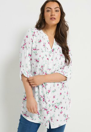 Kobieta o długich brązowych włosach, ubrana w białą kwiecistą koszulę na guziki z podwiniętymi rękawami i niebieskie jeansy, stojąca na jednolitym tle.