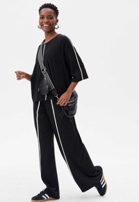 Zwarte oversized top en broek met brede pijpen met witte zijstrepen. Uitgerust met een zwarte crossbody tas en zwarte sneakers met witte accenten.