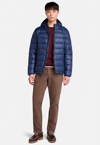 Marineblå dunjakke med vandrette sømme, lynlåslukning og justerbar hætte, båret over en kastanjebrun sweater, kombineret med brune bukser og snøresko.