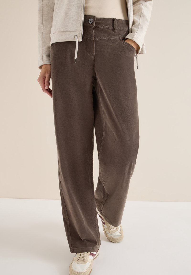 Personne portant un pantalon marron ample, une veste beige et des baskets blanches avec des accents bordeaux, debout devant un fond neutre.
