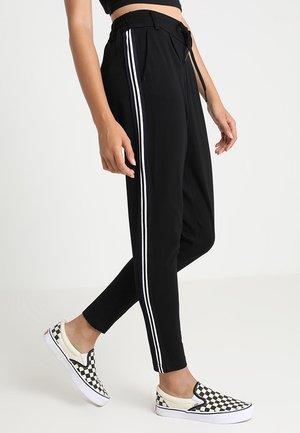 Person iført sorte tætsiddende bukser med hvide sidestropper og sort-hvide ternede slip-on sko, stående på et hvidt gulv.