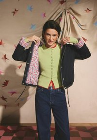 Jachetă bleumarin cu căptușeală florală, purtată peste un pulover verde cu nasturi și bordură roșie, combinată cu blugi albastri din denim, pe un fundal cu model.