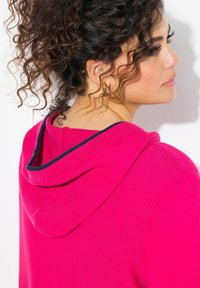 Różowa bluza z kapturem o teksturowanej fakturze, z granatowymi akcentami na kapturem. Charakteryzuje się luźnym krojem i okrągłym dekoltem z tyłu.