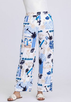 Frau trägt weiße Hosen mit weitem Bein und einem Muster aus blauen, beigen und schwarzen tropischen Motiven und Muscheln, kombiniert mit weißen Sandalen.