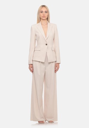 Donna in piedi che indossa un blazer beige chiaro su misura e pantaloni a gamba larga con sandali aperti su uno sfondo bianco uniforme.