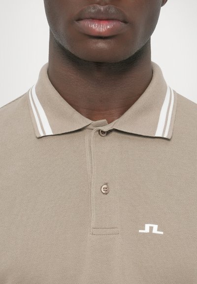 Μπεζ πόλο πουκάμισο με υφή, λευκές ρίγες στο γιακά και μικρό λευκό λογότυπο στο στήθος. Λεπτομέρεια με δύο κουμπιά στη λαιμόκοψη.