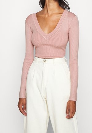 Femme portant un pull rose ajusté à côtes, manches longues avec col en V, et un pantalon blanc ample taille haute, debout devant un fond uni.