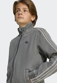 Veste zippée grise avec col montant, dotée de poignets et d'un ourlet côtelés, de trois bandes blanches sur les manches et d'un petit logo noir sur la poitrine.