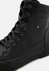 Svart høye sneakers har en teksturert skinnoverdel, svarte snører, sølvfarget øyelets og en gummisåle med et distinkt mønster.