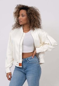 Veste bombardier blanche en satin avec des poignets côtelés, des poches zippées et une étiquette orange. Portée par-dessus un crop top blanc et un jean bleu.