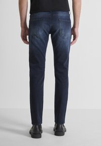 Jeans en denim bleu foncé avec une coupe droite, présentant des poches arrière et un léger délavage. Portés avec des bottes noires, vus de dos.