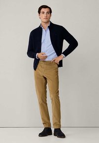 Άνδρας που στέκεται φορώντας μπλε ριγέ πουκάμισο, ναυτικό κασκόλ, καμηλό παντελόνι και μαύρα παπούτσια σε απλό φόντο.