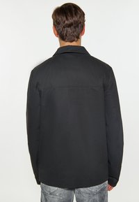 Svart jacka med krage, med en slät yta och rak skärning, sedd bakifrån, matchad med ljusgrå jeans.