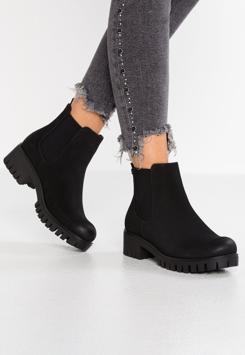 Personne portant des bottines noires à semelle épaisse associées à un jean skinny gris à ourlet effiloché avec des boutons décoratifs le long du côté.