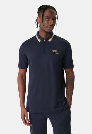 Uomo con capelli intrecciati che indossa una polo blu navy con colletto beige a righe e logo EA7 sul petto, in piedi con una mano in tasca.