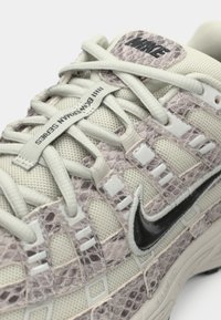 Λευκό και ροζ αθλητικό παπούτσι με υφή, επάνω μέρος από ύφασμα διαπνέον, διακοσμητικά που θυμίζουν ερπετό και μαύρο λογότυπο Nike στο πλάι. Διαθέτει λευκά κορδόνια και γλώσσα με το εμπορικό σήμα.