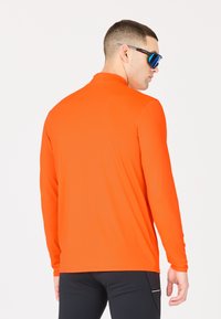 Långärmad idrottströja i stark orange, tillverkad av ett glättigt tyg. Har en hög krage och en figurnära passform, matchad med svarta idrottsbyxor.