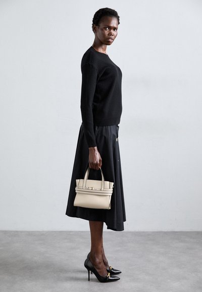 Femme en pull côtelé noir, jupe midi noire et escarpins noirs, tenant un petit sac à main beige, se tenant contre un mur clair.