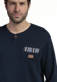 Camisa de manga larga navy con un bolsillo texturizado que presenta un patrón de pied de poule y una etiqueta marrón. Cuello redondo con cierre de botón.