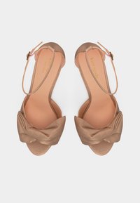 Γυναικεία μπεζ σανδάλια ανοιχτά στο μπροστινό μέρος με στριφογυριστά λουράκια και κλείσιμο με αγκράφα στον αστράγαλο, με επίπεδη σόλα, το όνομα της μάρκας τυπωμένο στο εσωτερικό.