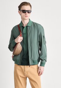 Zelená bomber bunda, zip vpředu, elastické manžety. Zelené polo tričko s logem. Béžové kalhoty. Hnědá potisková kabelka přes rameno. Černé sluneční brýle.