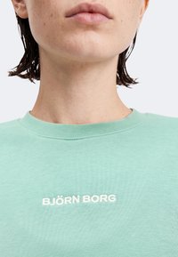 Άτομο που φοράει ανοιχτοπράσινο μπλουζάκι με στρογγυλή λαιμόκοψη και λευκό κείμενο "BJÖRN BORG" στο κέντρο του στήθους, ορατό από το σαγόνι μέχρι τον πάνω κορμό.