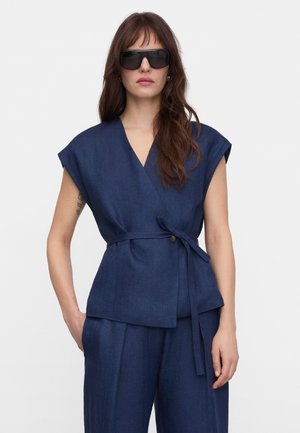 Femme portant un haut cache-cœur sans manches bleu foncé avec ceinture à nouer et un pantalon large assorti, aux longs cheveux bruns et portant de grandes lunettes de soleil noires.