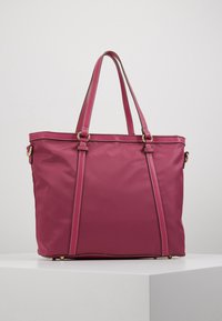 Duża różowa torba na ramię z podwójnymi paskami, złotymi akcentami i białymi przeszyciami, umieszczona na białej powierzchni na tle w neutralnych kolorach.