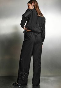 Schwarzes Zweiteiler-Outfit mit einem Shirt mit floralem Stickereimuster und lockeren Hosen. Der Stoff ist glatt und hat ein mattes Finish.