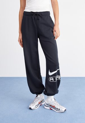 Persona che indossa pantaloni della tuta Nike neri con logo bianco e scarpe da ginnastica bianche, in piedi su un pavimento blu contro una parete bianca.