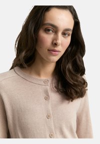 Frau mit langen, dunklen, lockigen Haaren, die einen beigen, geknöpften Cardigan trägt und leicht zur Seite blickt, vor einem schlichten Hintergrund.