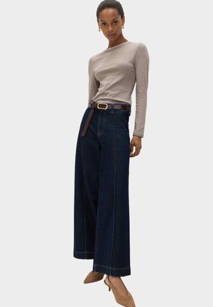 Mujer con camiseta de manga larga beige, vaqueros anchos oscuros con cinturón marrón y zapatos de tacón marrón puntiagudos, de pie frente a un fondo claro y liso.