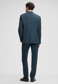 Man in een donkere teal suit en zwarte geklede schoenen staat met zijn rug naar de camera tegen een effen lichtgrijze achtergrond.