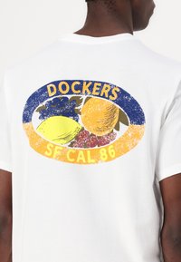 Hvit bomull t-skjorte med et stort, fargerikt grafikk av frukter og teksten "DOCKERS SF CAL 86" i blått og gult, med en slitt finish.