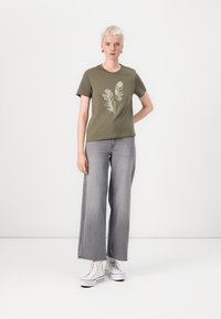 Maslinasto zeleni T-shirt s printom perja, u kombinaciji sa širokim sivosivim trapericama. Model nosi bijele visoke tenisice, s kratkom svijetlom kosom i naušnicama.