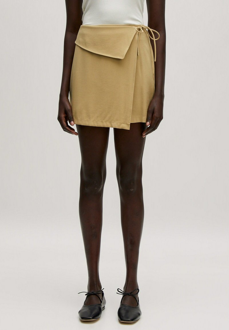 Khaki wikkelrok met een gestrikte taille, voorzien van een klepzak en zachte, gladde stof. Gemaakt met zwarte flats en doorschijnende tights.