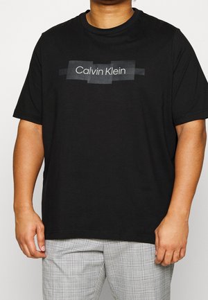 Černé bavlněné tričko s jednoduchou grafikou obsahující "Calvin Klein" v bílo-šedém lineárním designu. Modelka má na sobě kostkované šedé kalhoty.