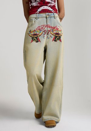 Bredbenede, falmede jeans med farverigt oksehoveddesign og teksten "Ed Hardy" på baglommerne, båret med beige støvler og en rød grafisk top.