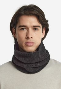 Sciarpa con collo a scialle lavorata a maglia di colore grigio scuro, con un motivo testurizzato e dettagli a costine, indossata sopra un maglione chiaro.