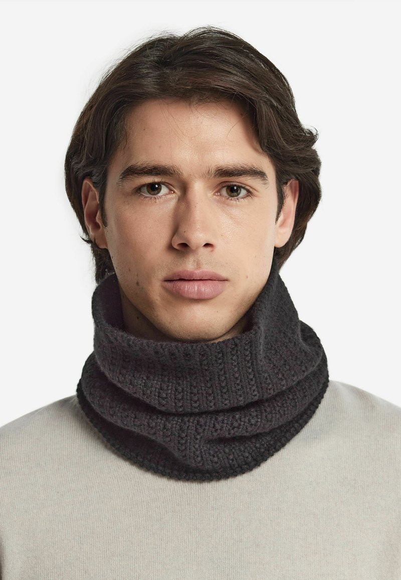 Sciarpa con collo a scialle lavorata a maglia di colore grigio scuro, con un motivo testurizzato e dettagli a costine, indossata sopra un maglione chiaro.