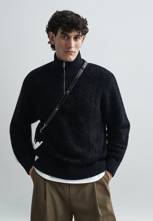Sweater preto de malha com gola alta, superfície texturizada e punhos canelados. Combinado com calças pregueadas em tom areia e uma bolsa tiracolo preta.