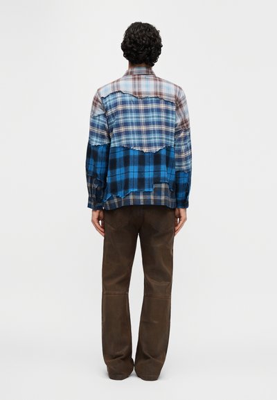 Καρό πουκάμισο φανέλας με μπλε, καφέ και γκρι τμήματα, διαθέτει ακανόνιστο τελείωμα, μακριά μανίκια και άνετη εφαρμογή. Συνδυασμένο με καφέ παντελόνια.