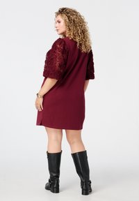 Robe bordeaux avec des manches transparentes brodées et texturées, longueur genoux, fentes sur les côtés, associée à des bottes noires hautes pour un look audacieux.