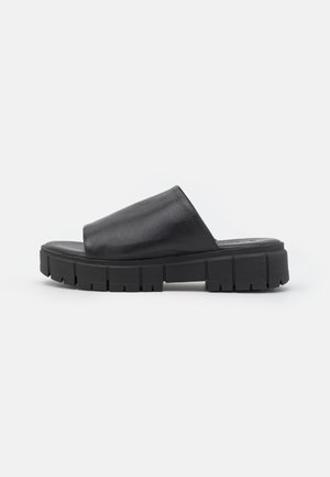 Zwarte slipper met brede leren band en dikke, grove rubberen zool met diepe profielpatronen, zijaanzicht op een effen achtergrond.