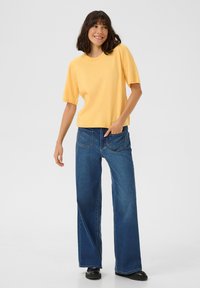 Un haut jaune en tricot à manches courtes associé à un jean en denim bleu à jambes larges et des chaussures noires. Le haut a un col rond et un tissu légèrement texturé.
