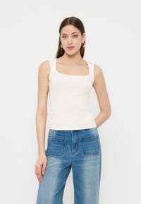 Femme aux longs cheveux bruns portant des boucles d’oreilles créoles dorées, un top blanc sans manches à encolure carrée et un jean bleu clair à jambes larges.