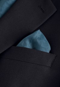 Veste de costume texturée sombre avec un carré de poche bleu à motif plié glissé dans la poche poitrine.