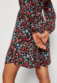 Schwarzes Kleid mit floralem Muster, das rote, rosa und blaue Blumen zeigt. Lange Ärmel, tailliert und mit abgerundetem Saum. Glatte Stoffstruktur.