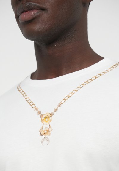 Zlatý náhrdelník s kvetinovými akcentmi a detailom polmesiaca, nosený na bielej košeli. Textúrované kovové očká vytvárajú kontrast.